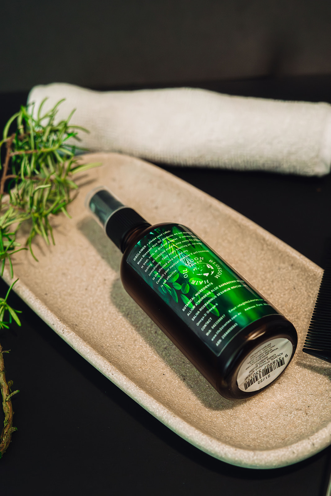 Moon Blossom Hair Oil bottle resting on a stone tray with rosemary and a towel